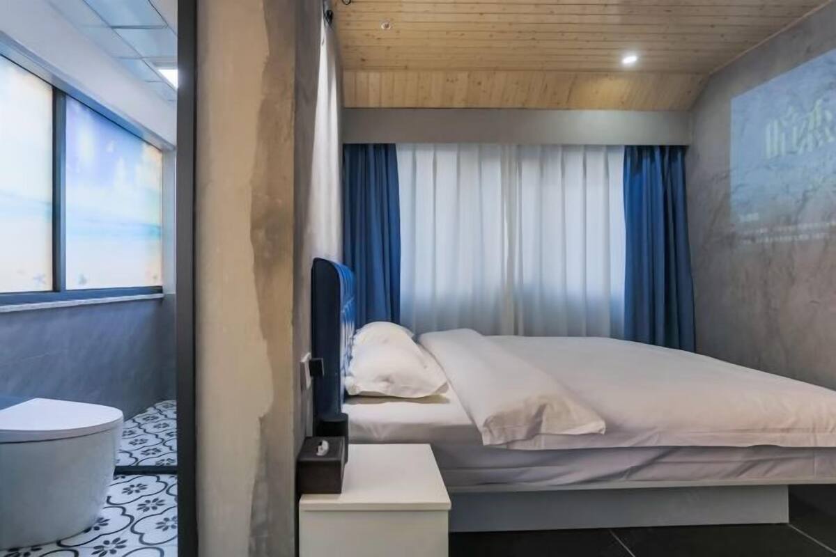 A hotel room with a bed, a bedside table, a window with curtains, and a bathroom with a toilet and patterned floor.