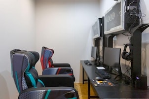 Uma cadeira gamer com padrão geométrico, uma escrivaninha com computador, incluindo monitor, teclado e rato, e uma TV fixada na parede.