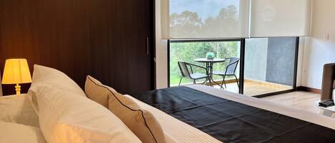 A hotel room with a bed, a nightstand with a lamp, a balcony with a table and chairs, and a view of trees.