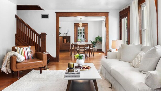A living room with a brown leather sofa, a white sofa, a coffee table with books and a plant, a wooden staircase, and a dining area with a table and chairs. - Stylish Historic Home – Timeless Charm with Modern Touches (Bovina Center)