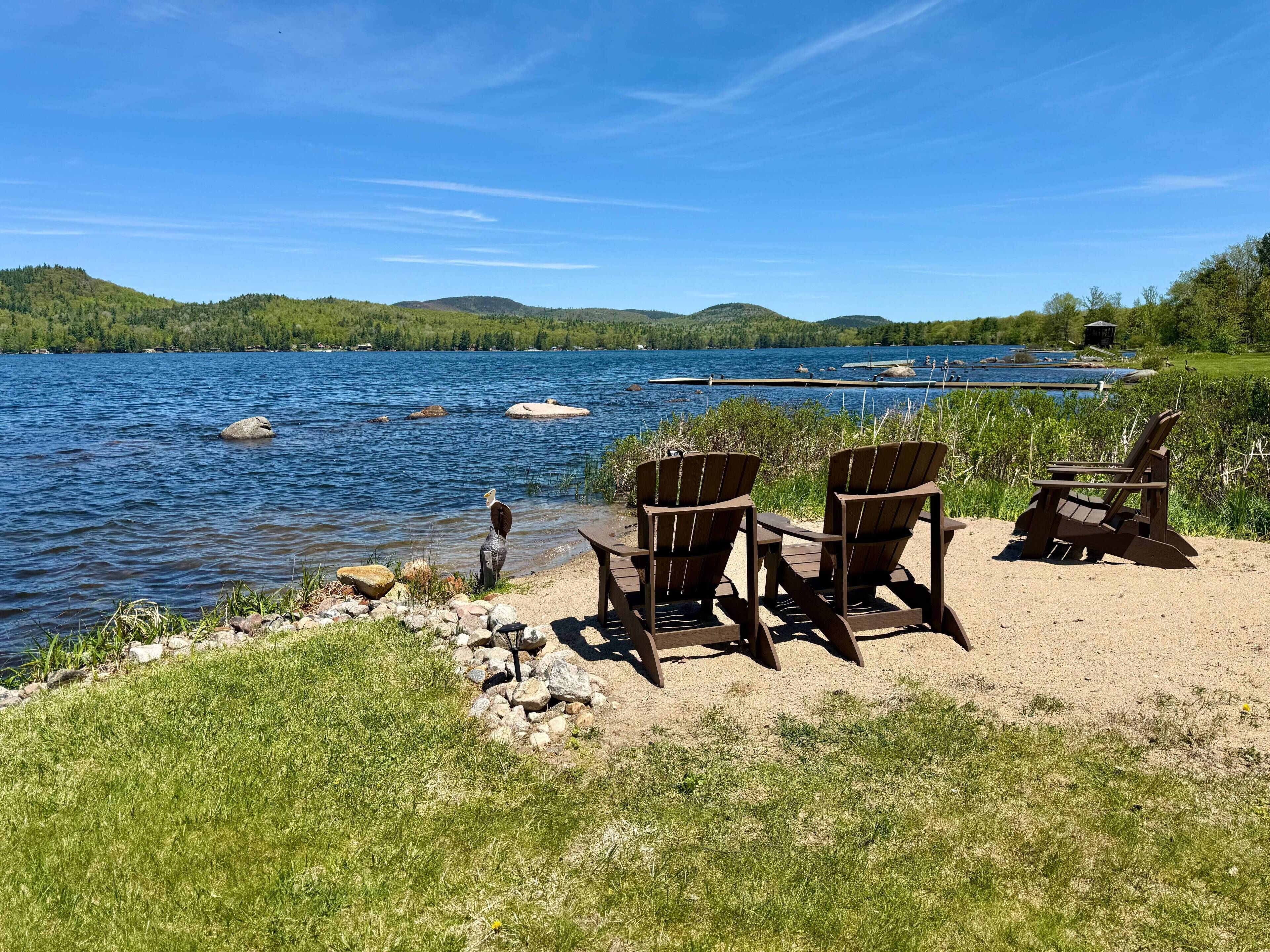A lakeside view with chairs and a table, surrounded by greenery and hills.