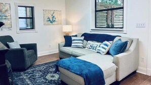 Un salon avec un canapé, une table basse et un tapis. Il y a deux fenêtres et un tableau au mur.