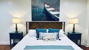 Une chambre à coucher bien rangée, avec un lit placé au centre, entre deux tables de chevet, chacune surmontée d’une lampe. Au-dessus du lit, on peut voir un tableau représentant un bateau.