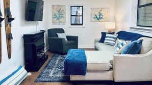 Un salon comprenant un canapé, un fauteuil et un téléviseur fixé au mur. Deux tableaux ornent le mur, et une fenêtre offre une vue agréable.