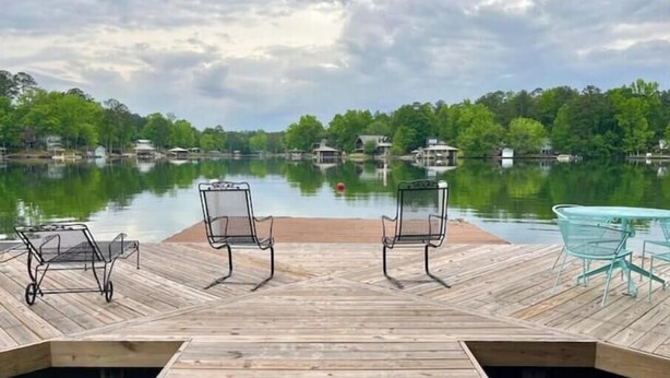 Une jetée en bois avec deux chaises et une table, d’où l’on a vue sur un lac paisible, avec des maisons en arrière-plan.