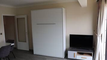 Ein Zimmer mit einem großen, weißen Kleiderschrank, einem Flachbildfernseher auf einem Schrank und einem Esstisch mit Stühlen. Eine Tür führt in einen weiteren Raum.