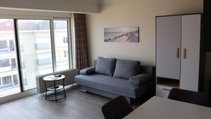 Un salon moderne avec un canapé gris, une petite table ronde et une table à manger. Une grande fenêtre offre une vue sur les bâtiments environnants.