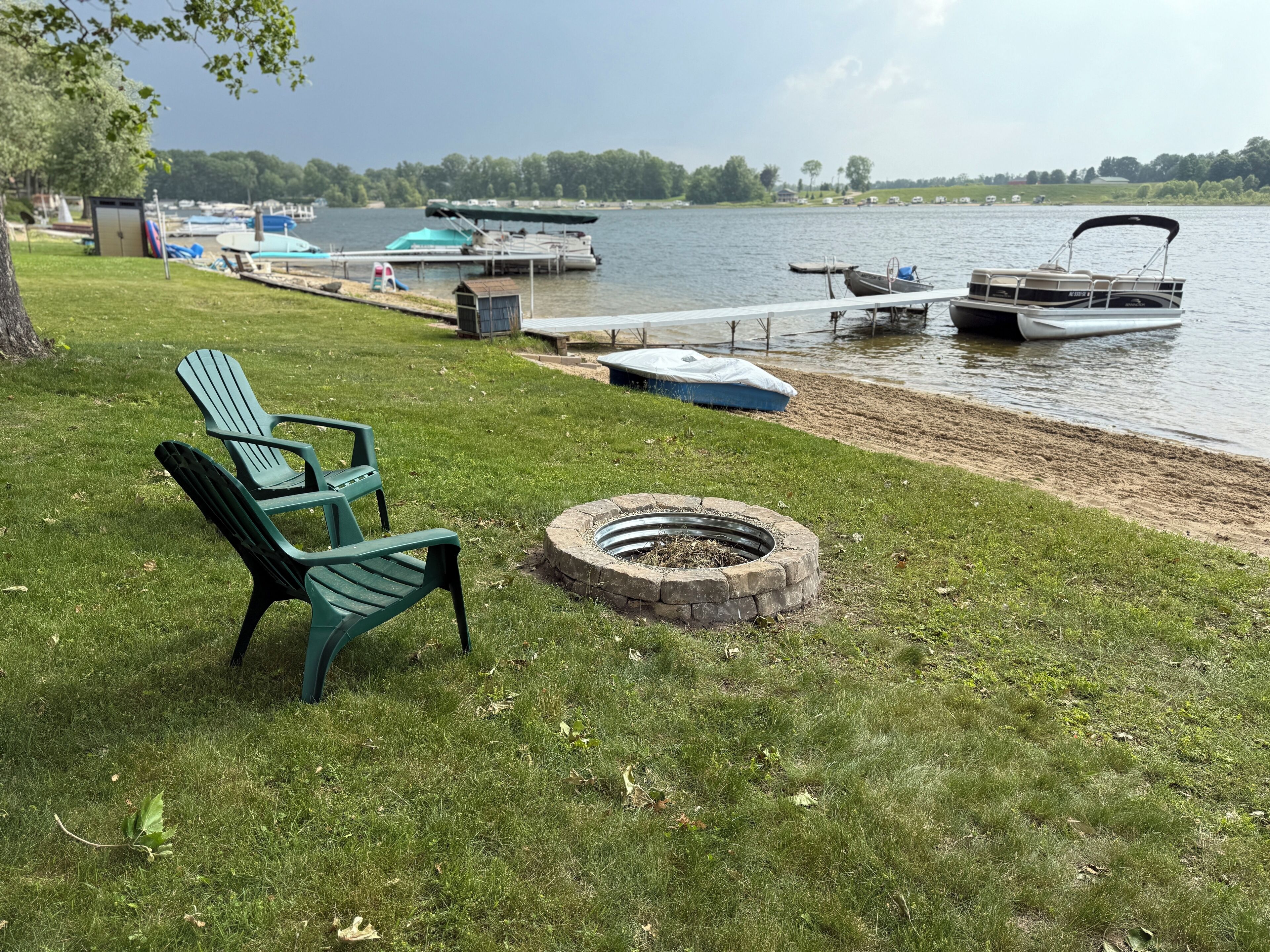 A lakeside deck with a fire pit, a green Adirondack chair, and a dock with boats.