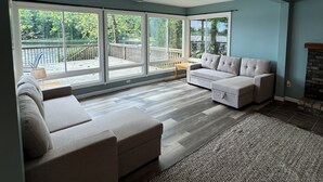 A spacious living room with a large sectional sofa, a fireplace, and a view of a deck and trees. - Charming Lakefront Retreat with Sandy Beach – Perfect Getaway  (Shelbyville)