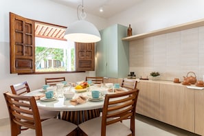 Un comedor con una mesa redonda, sillas de madera, una lámpara colgante y vistas al exterior a través de una ventana con contraventanas de madera.