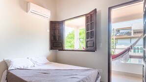 A bedroom with a bed, an open window, a hammock outside, and an air conditioning unit on the wall.