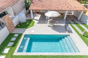 Une piscine avec une terrasse carrelée, un espace de détente couvert avec des chaises et un parasol, et un mur en briques.