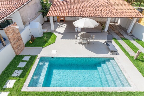 A swimming pool with a tiled deck, a covered seating area with chairs and an umbrella, and a brick wall. - Cassa em Cond beira-mar-4qts-SH088B (Tamandaré)