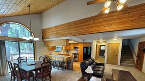 A spacious living area with a dining table, chairs, a leather sofa, and a coffee table. A kitchen with wooden cabinets and appliances is visible in the background.