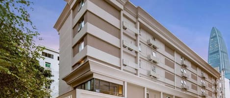 Um hotel com vários andares, fachada moderna, varandas e um edifício de vidro imponente ao fundo.