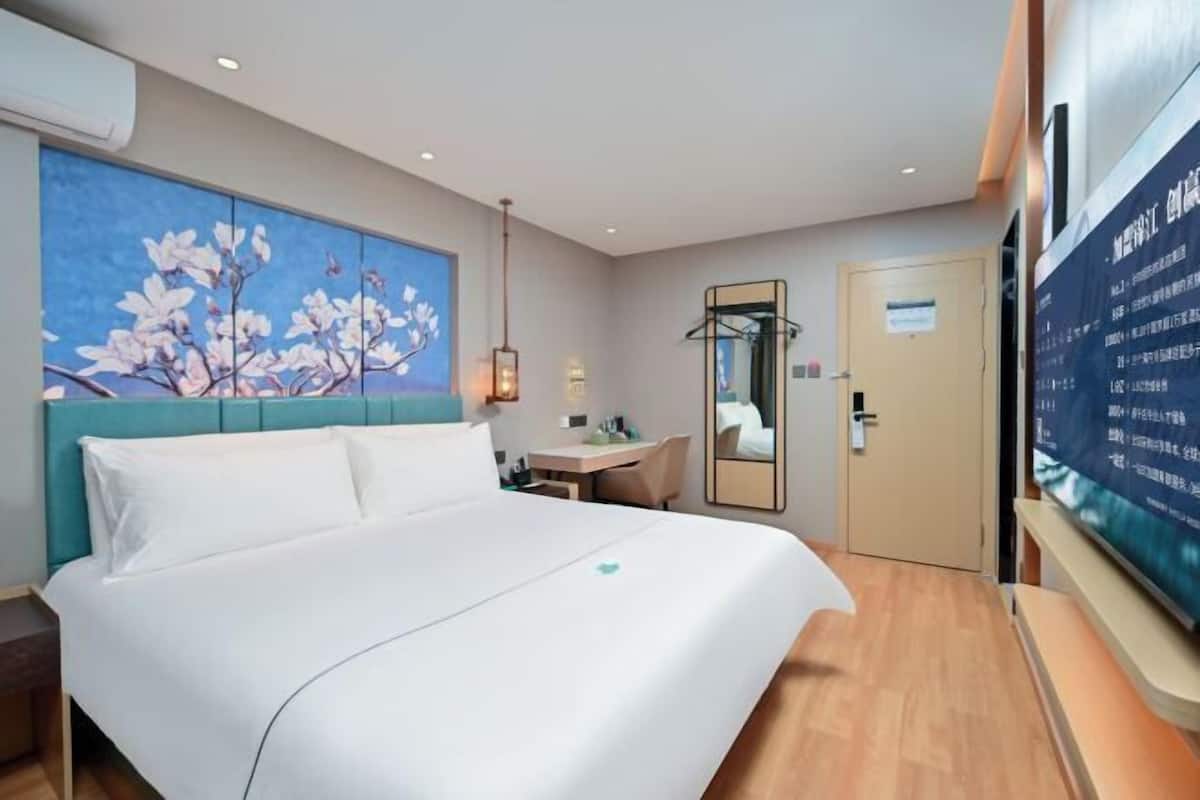 A hotel room with a bed, a desk, a chair, a mirror, and a wall-mounted TV.