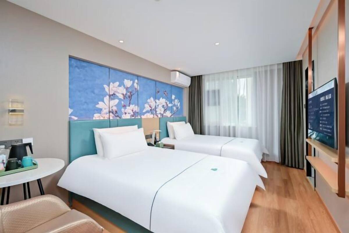 A hotel room with two beds, a desk with a coffee maker, a TV, and a large floral artwork on the wall.