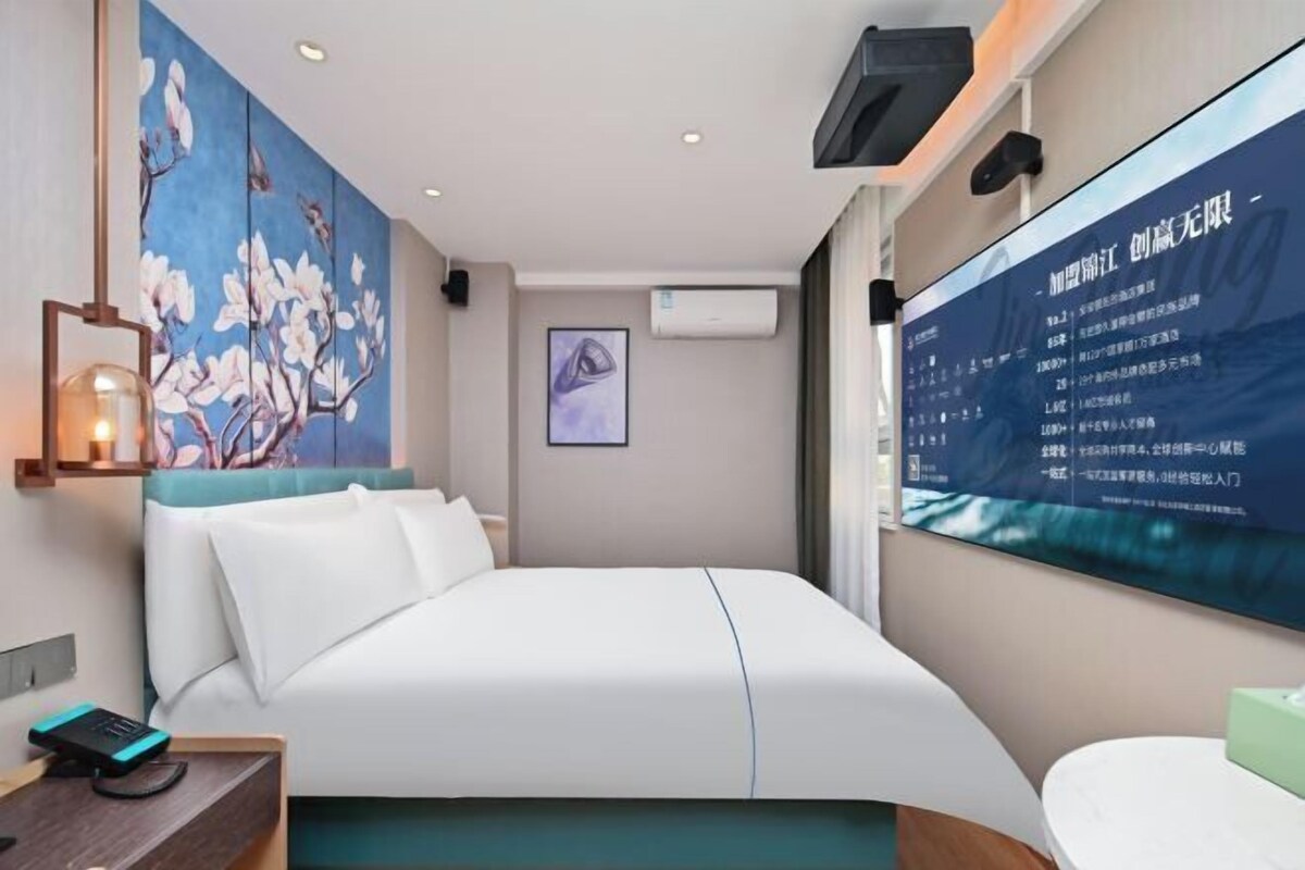 A modern hotel room with a large bed, a desk, and a wall-mounted TV displaying a menu.