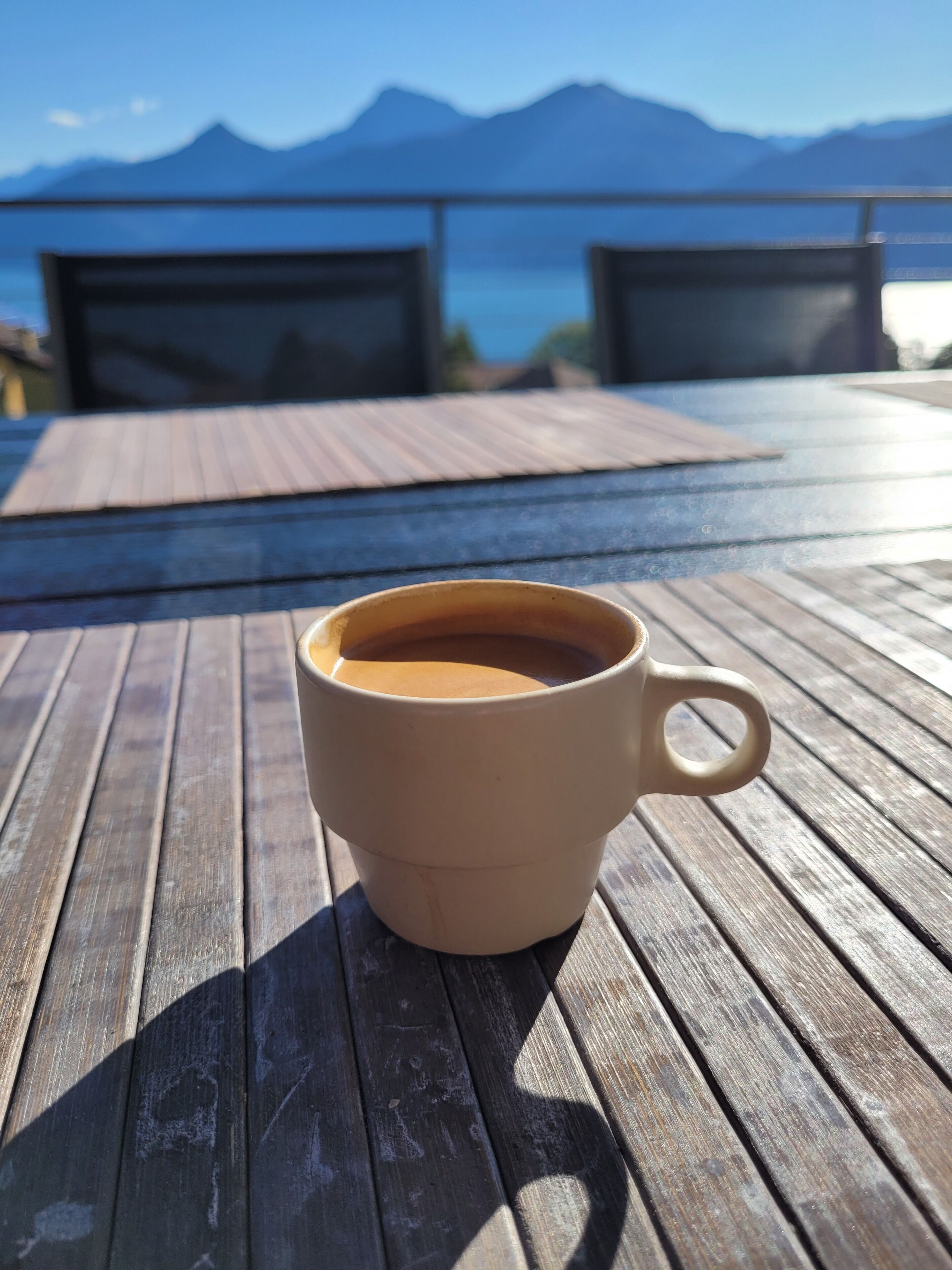 A cup of coffee on a wooden deck with a view of mountains and a body of water in the background.