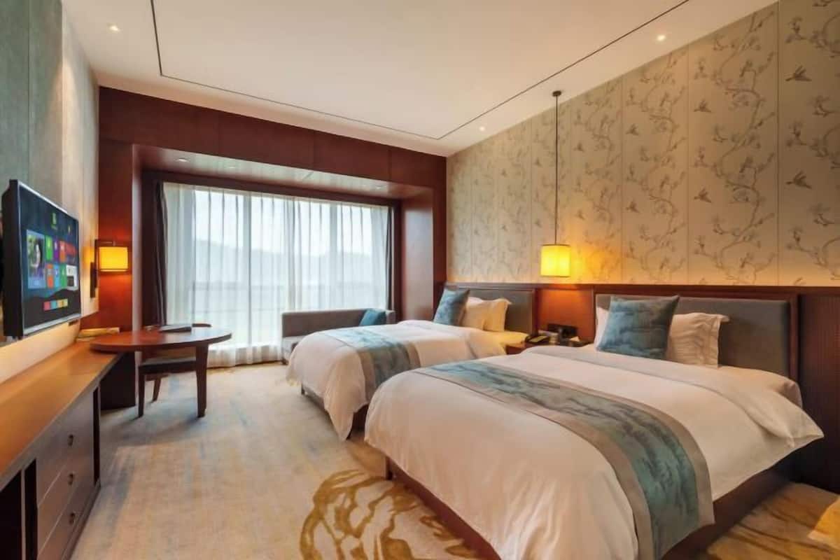 A hotel room with two beds, a TV, a desk, and a window with curtains.