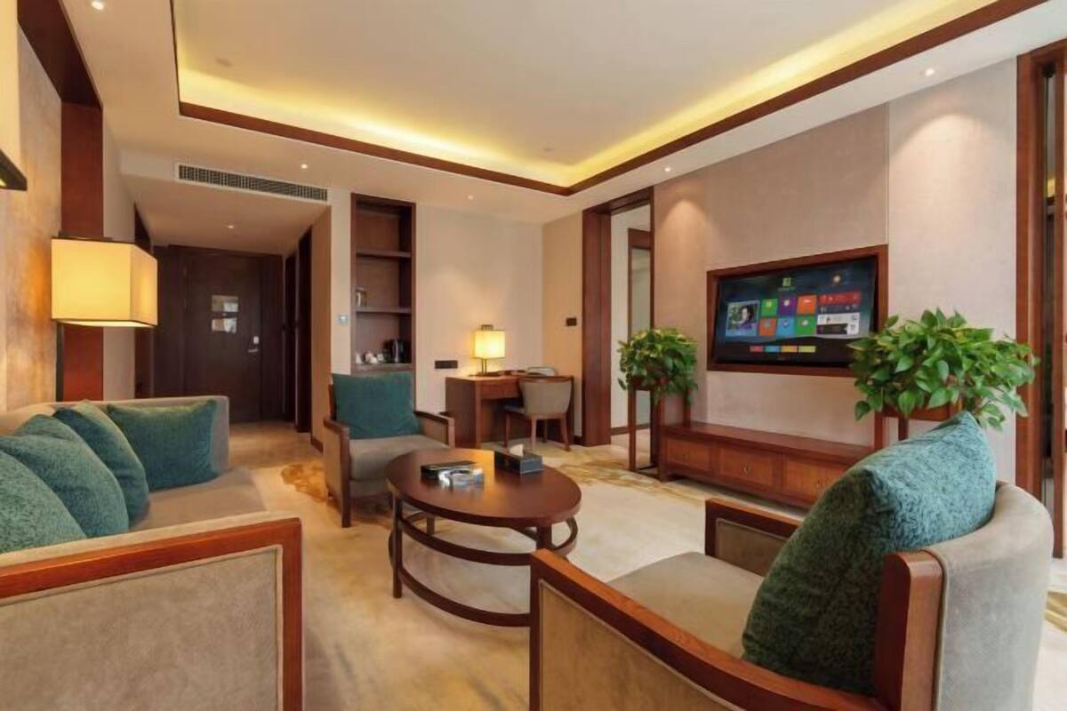 A hotel room with a sofa, armchair, round table, TV, and two potted plants.