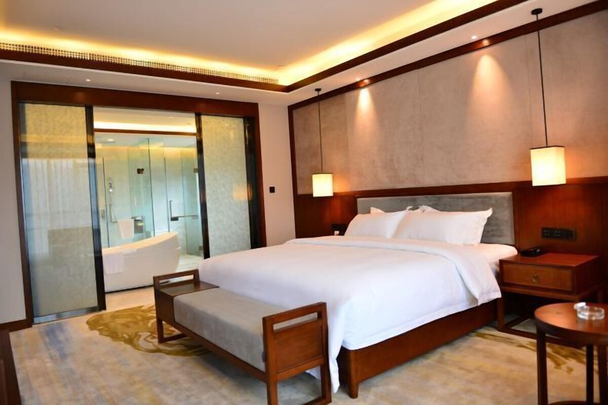 A hotel room with a large bed, a bedside table, a nightstand, a wooden chair, a small round table, a glass-enclosed bathroom with a bathtub, and a large mirror.