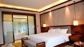 A hotel room with a large bed, a bedside table, a nightstand, a wooden chair, a small round table, a glass-enclosed bathroom with a bathtub, and a large mirror.