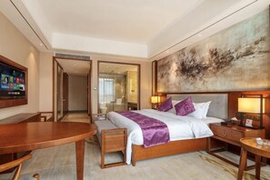 A hotel room with a bed, bedside table, lamp, TV, and a large abstract painting on the wall.