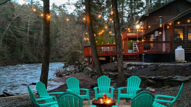 Une cabane dotée d’un foyer extérieur et de sièges, entourée d’une forêt et bordée par une rivière.