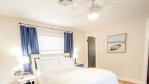 Une chambre à coucher avec un lit, une table de chevet, un ventilateur de plafond, une fenêtre avec des rideaux et un tableau accroché au mur.