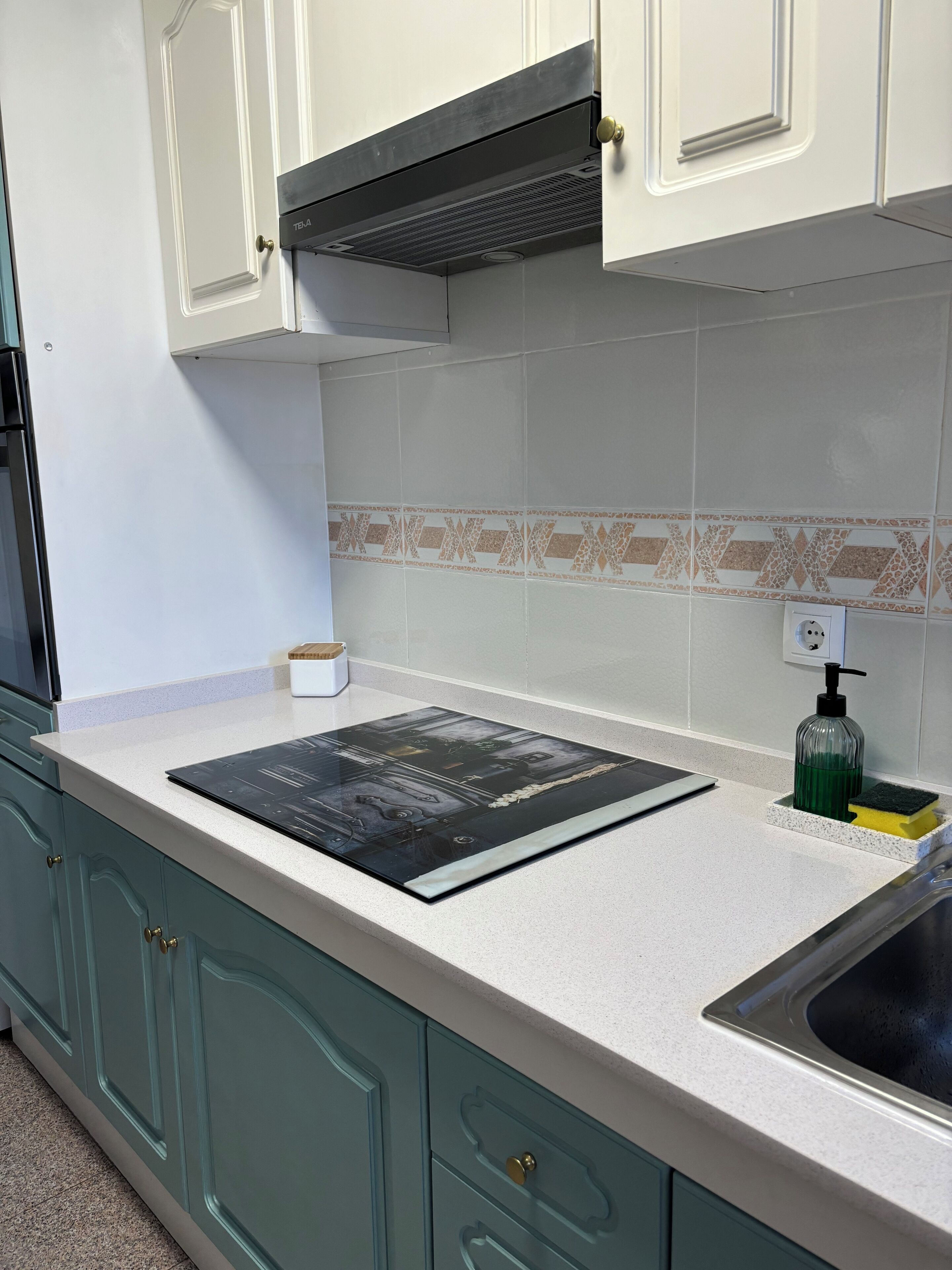Una cocina con armarios color turquesa, una encimera blanca, una cocina por inducción negra y un fregadero de acero inoxidable.