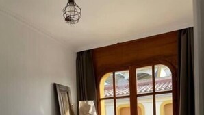 A bedroom with a large bed, a chair, a table, and a window with a view of a building with a tiled roof. - Calafell Beachfront Escape. Spacious Attic with Rooftop Terrace and Pool. (CT)