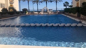 A swimming pool with a clear view of the ocean in the background, surrounded by palm trees and a building with balconies. - Calafell Beachfront Escape. Spacious Attic with Rooftop Terrace and Pool. (CT)