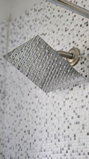 A showerhead with a metallic finish and a square design, mounted on a tiled wall with a mosaic pattern.