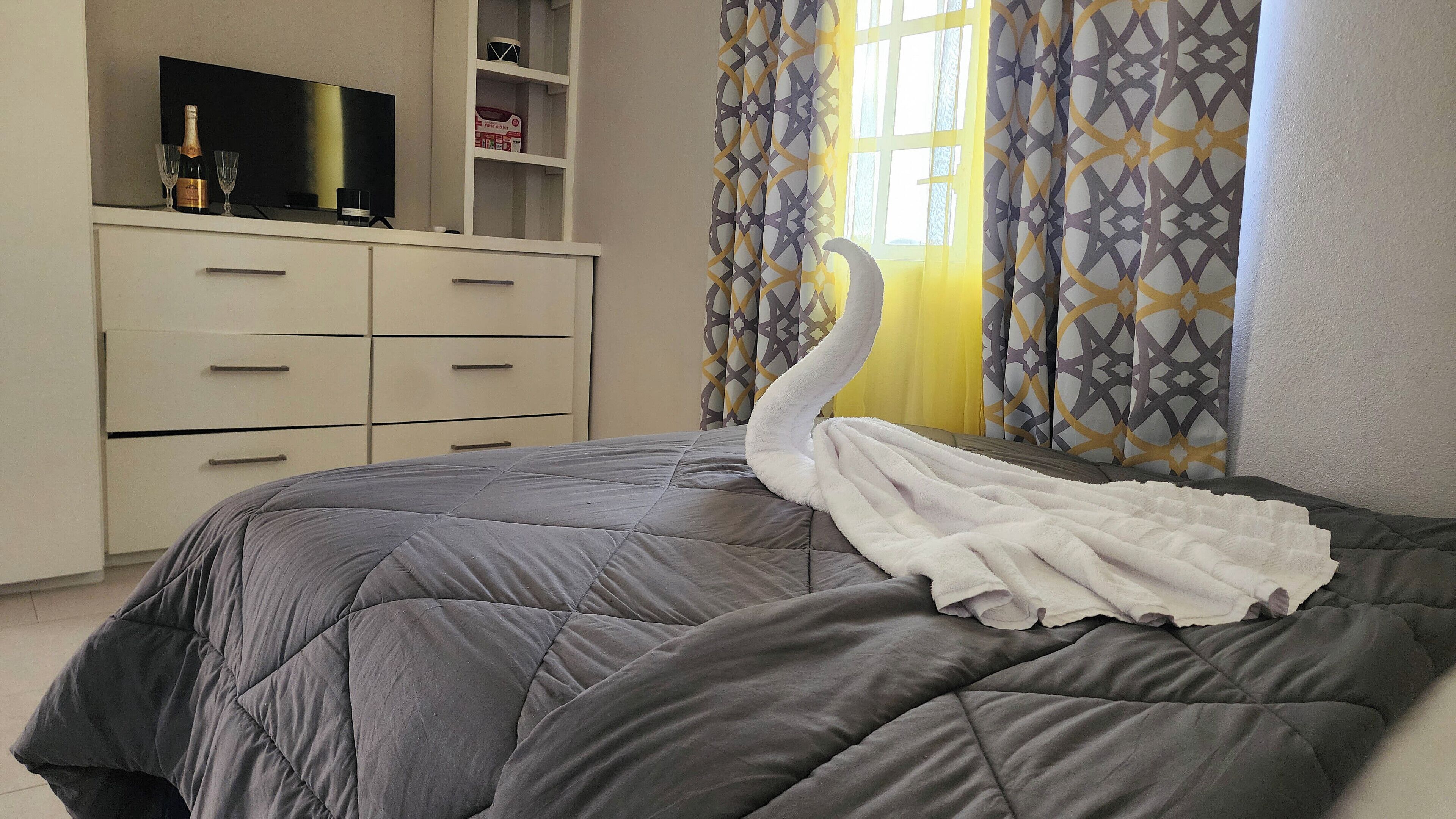 A bedroom with a bed, a dresser, a TV, and a window with patterned curtains.