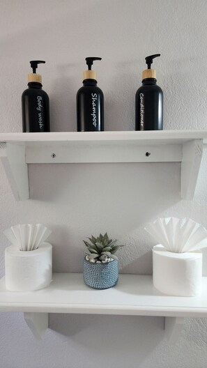 A white shelf with three black bottles labeled "Body wash," "Shampoo," and "Conditioner," and two white tissue rolls with a small potted plant in between.