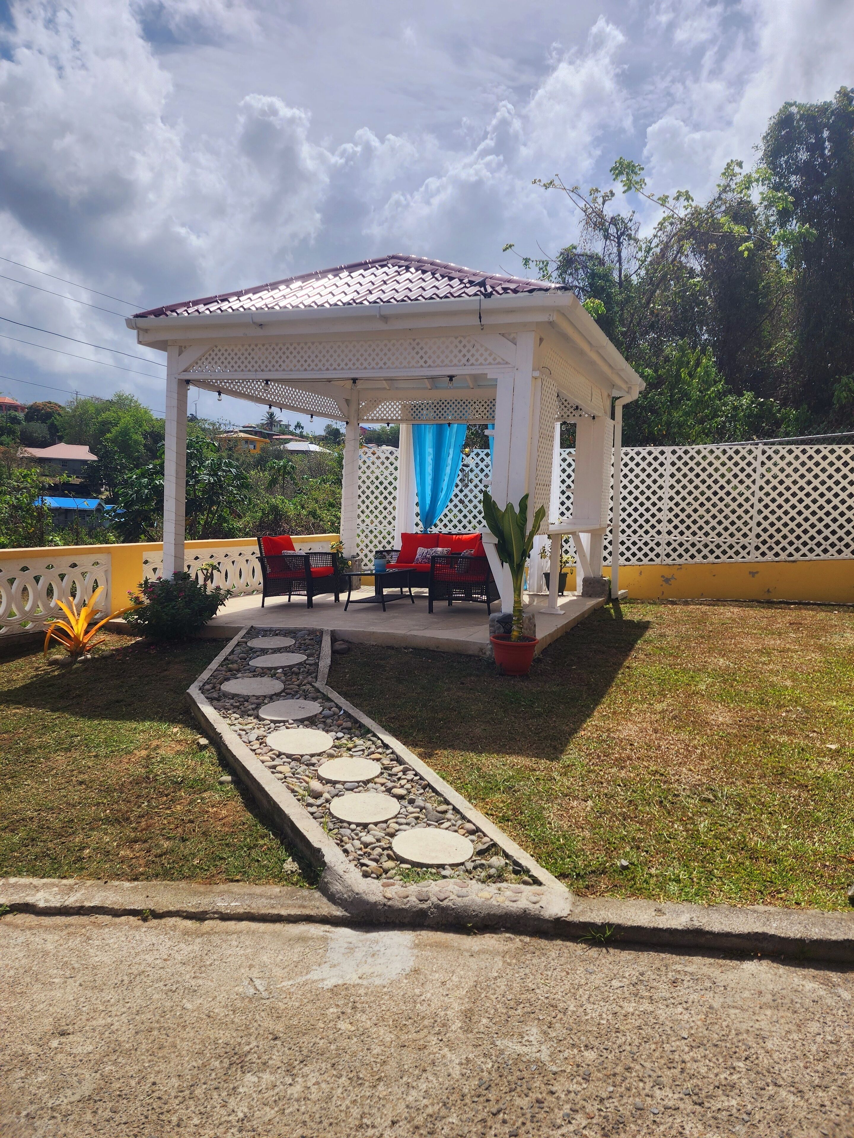 Uma área de estar ao ar livre coberta com telhado de azulejos, rodeada por uma cerca branca, e um caminho que a leva.