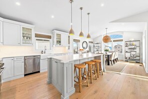Una cocina amplia con alacenas blancas, una isla grande con taburetes de bar, iluminación colgante y vistas al área de comedor.