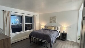 Un dormitorio con cama, mesita de noche, ventana con cortinas e iluminación en la pared.
