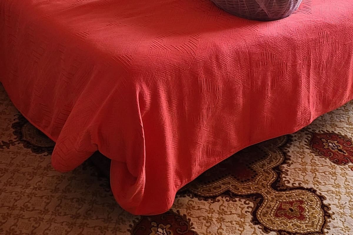 Un lit bien fait, recouvert d’une couverture rouge, un panier en osier rempli de serviettes, une table de chevet avec une lampe et un tableau encadré accroché au mur.