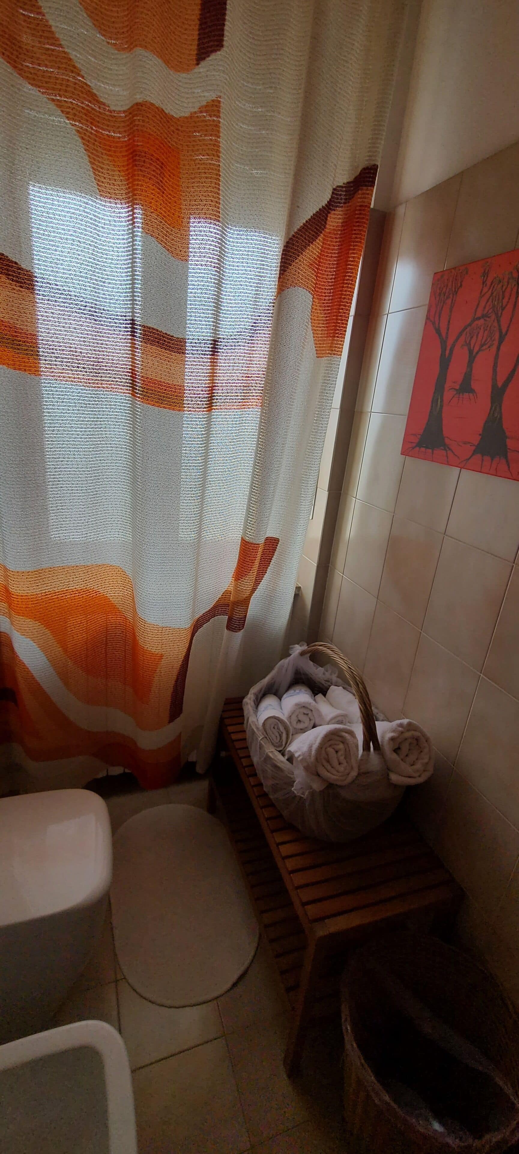Un baño con cortina de ducha, un taburete de madera con toallas, un inodoro y un cubo de basura.