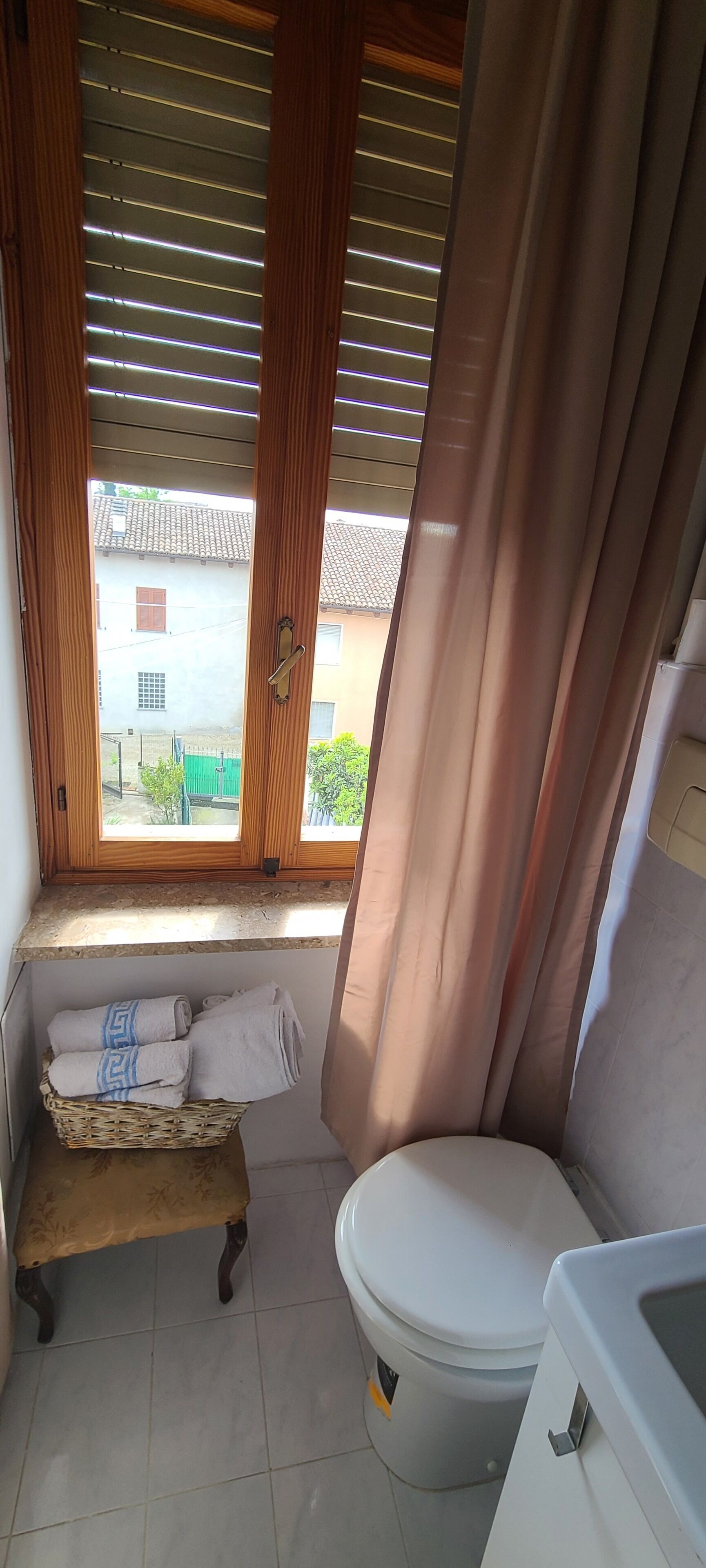 Et badeværelse med toilet, vask, et vindue med træpersienner og en lille bænk med håndklæder.