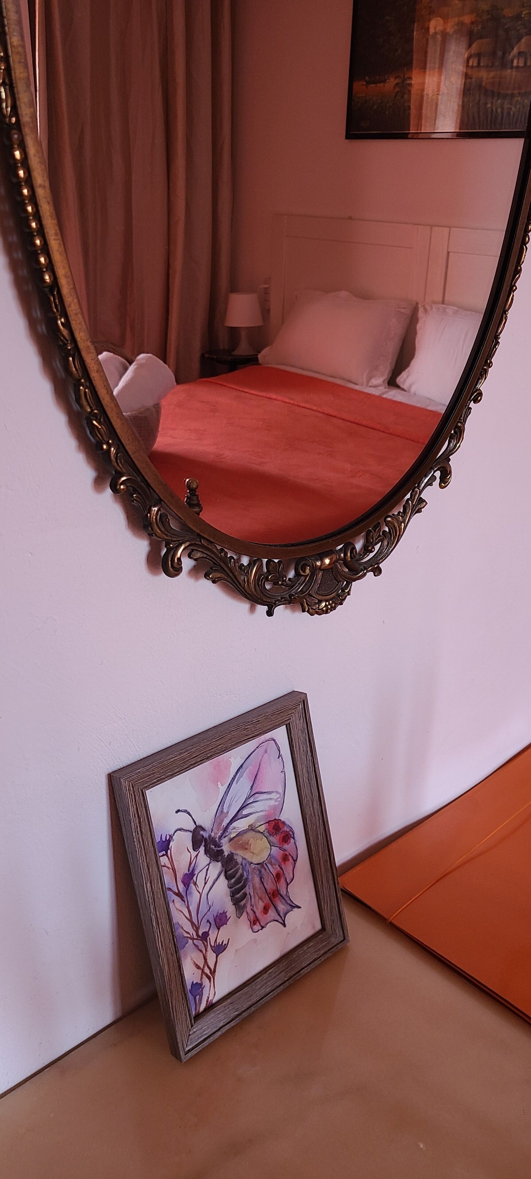 Une chambre à coucher avec un grand miroir décoratif, un lit recouvert d’une couverture rouge et un tableau encadré représentant un papillon accroché au mur.