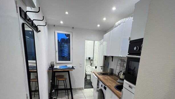 A compact kitchen with a washing machine, a dining table with chairs, and a view of the outside through a window.