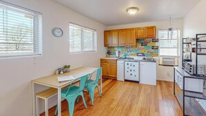 A compact kitchen with wooden cabinets, a white countertop, and a small island. There are two windows with blinds, a clock on the wall, and a ceiling light fixture.