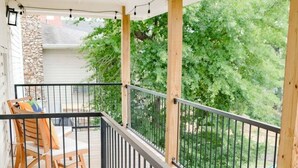 Un terrazza in legno a più livelli con ringhiera in metallo, una zona salotto coperta con sedie e vista su un muro in pietra e vegetazione.