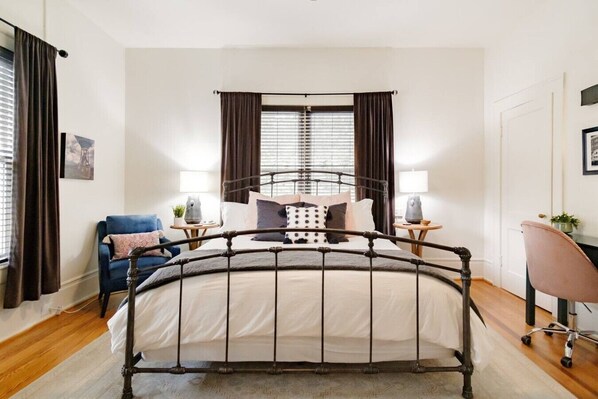 Un dormitorio con una cama de estructura metálica, dos mesitas de noche con lámparas, un sillón azul, un escritorio con silla y una ventana con persianas.