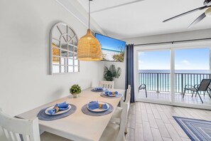 Un comedor con una mesa dispuesta para dos personas, una lámpara colgante, una planta en maceta y vistas al océano a través de un ventanal corredera.