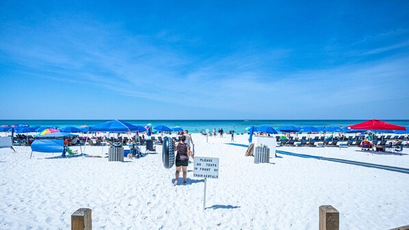 A beach with white sand, blue umbrellas, and a sign prohibiting tents near the chair rentals.