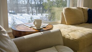 Ein gemütliches Zimmer mit Blick auf eine verschneite Landschaft durch ein Fenster, ein runder Holztisch mit Tasse und Speisekarte sowie ein beiges Sofa mit Kissen.